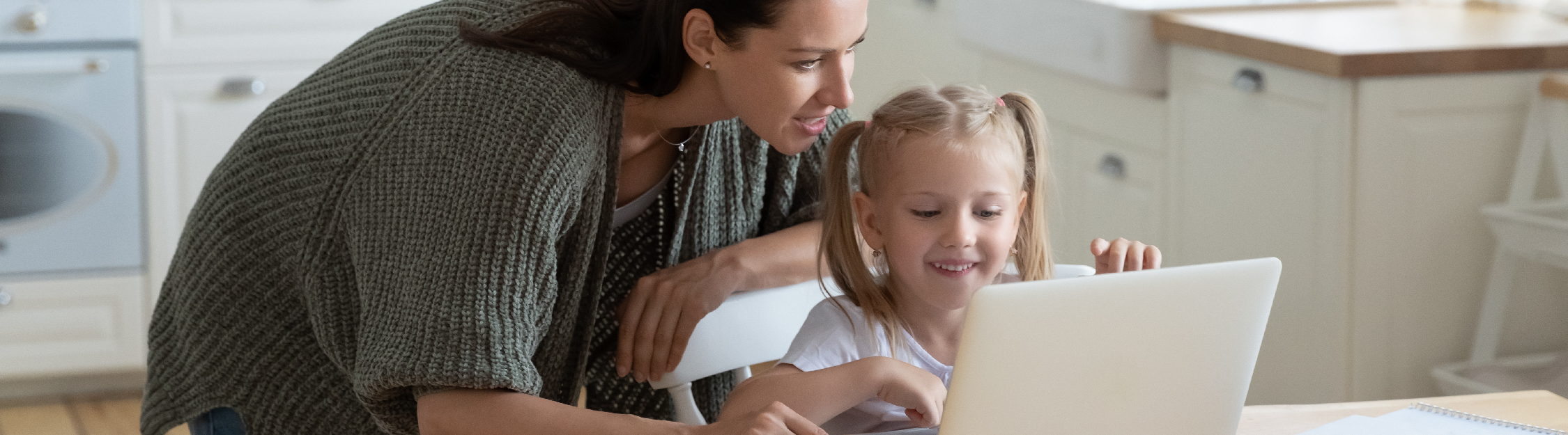 Mother and toddler daughter smile while using laptop in kitchen